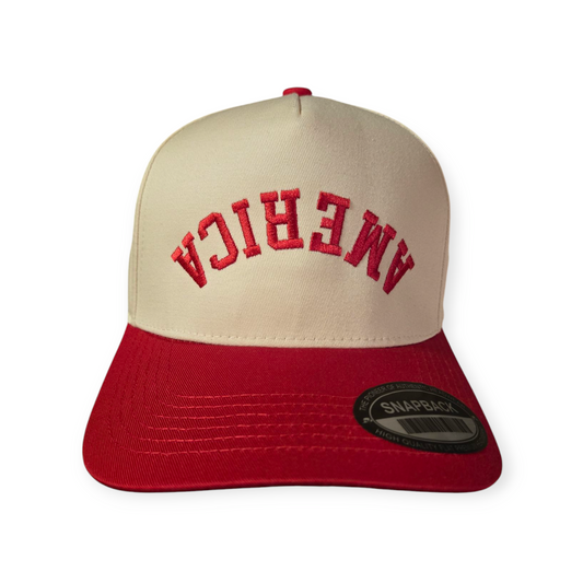 Front facing America Upside Down V1 structured hat in red and beige with the word “America” embroidered upside down in red on the front.