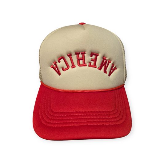 Front facing America Upside Down V1 trucker hat in red and beige with the word “America” embroidered upside down in red on the front.