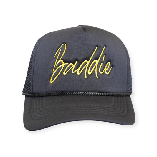 Front-facing black Baddie trucker hat with a curved brim and mesh back, featuring “Baddie” embroidered in gold across the front.